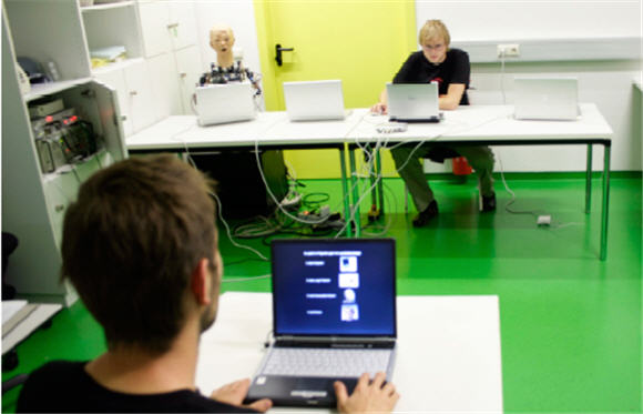 Un cobaye humain (de dos) face à ses 4 partenaires : le robot humanoïde, le bras robotique, un être humain et le programme informatique,